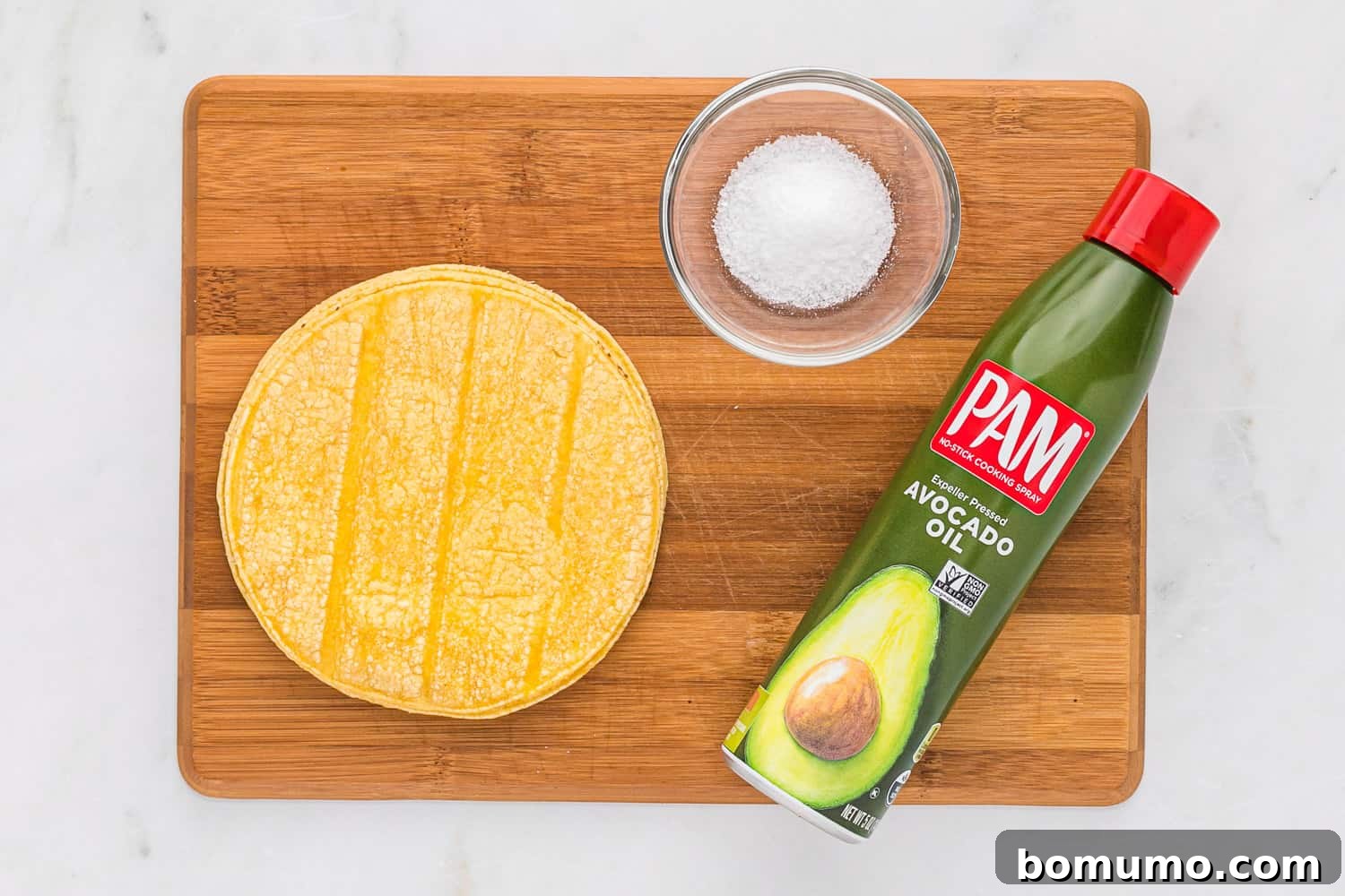 Crispy Oven Tortillas 4 A wooden cutting board displaying a stack of corn tortillas, a bottle of avocado oil spray, and a small pile of kosher salt, the three main ingredients for baked tortilla chips.