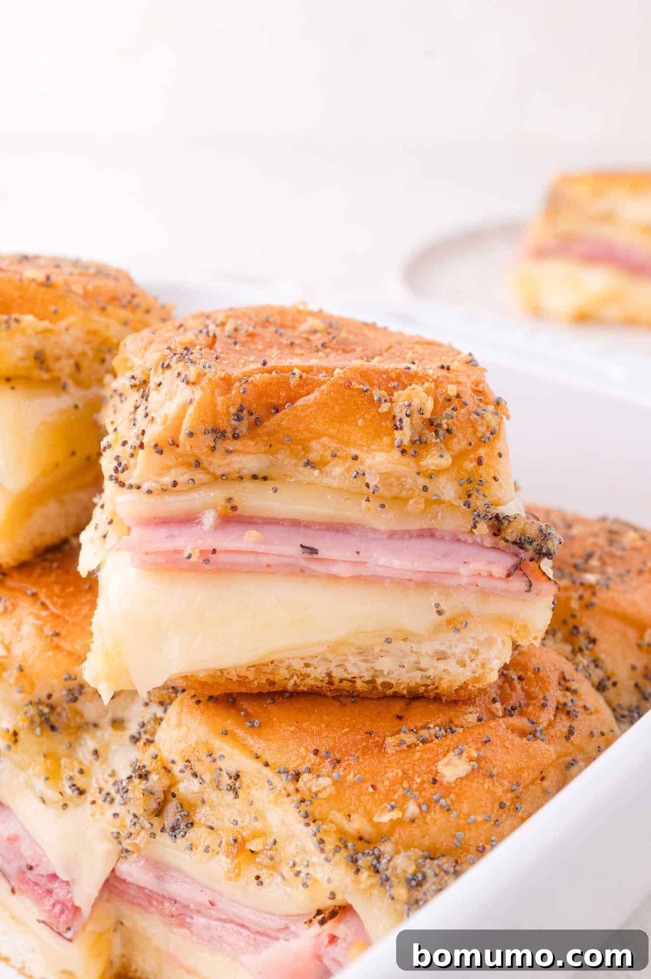Close-up of baked ham and cheese sliders with poppy seeds on top, served in a white dish.