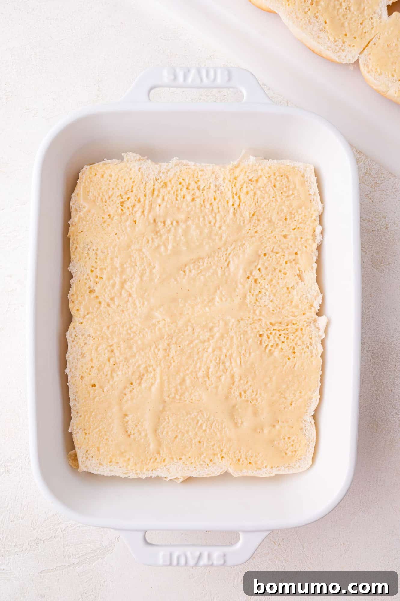 A white baking dish containing a single layer of pale, spongy rolls, topped with mayo and mustard mixture.
