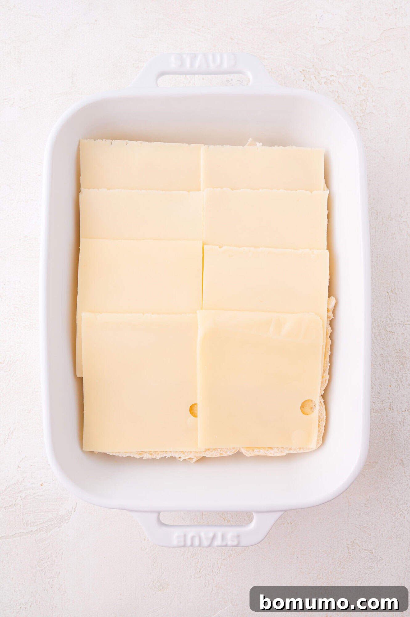 A white baking dish with neatly arranged slices of Swiss cheese covering the rolls on the bottom.