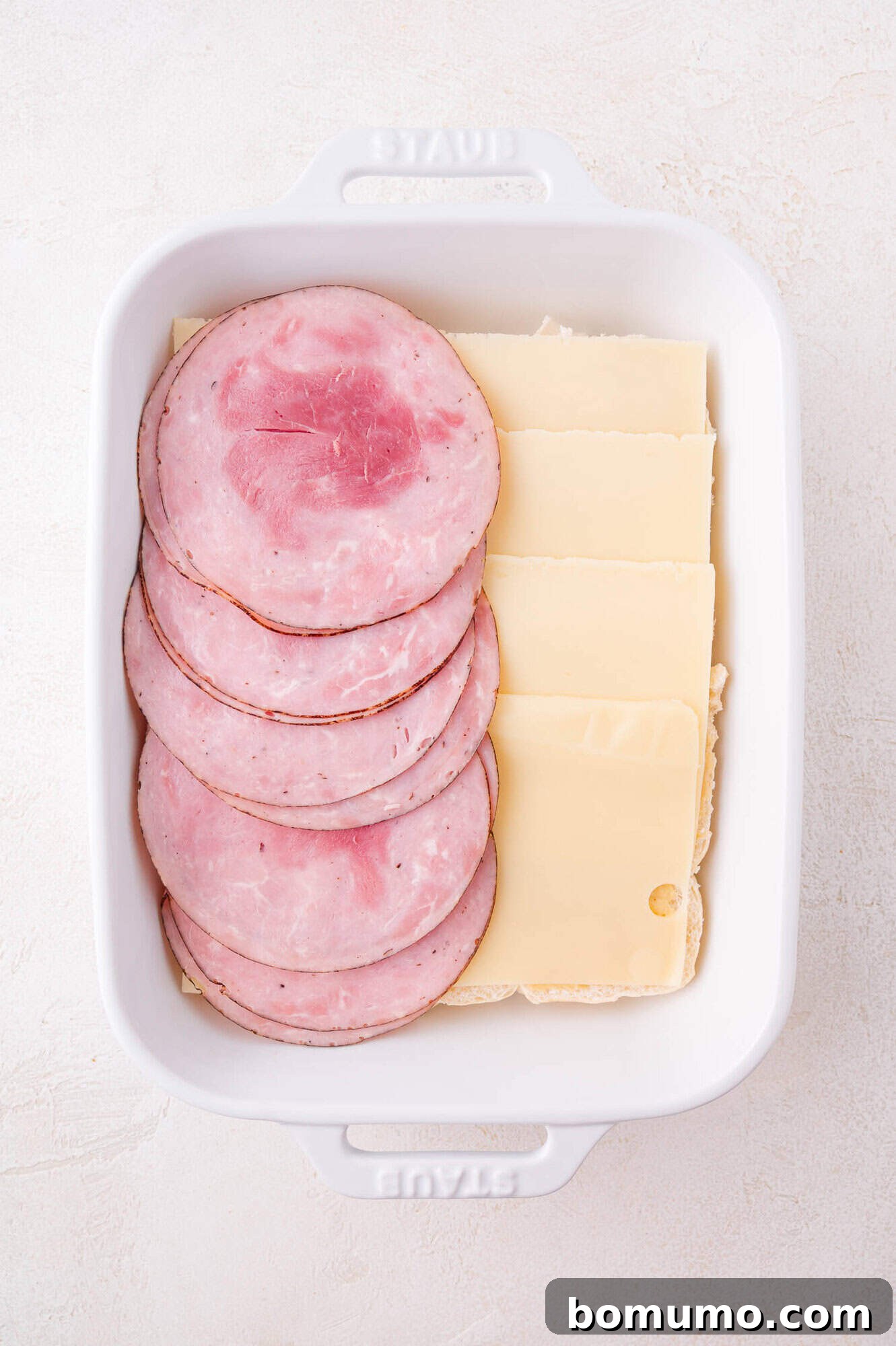 A white baking dish containing overlapping slices of ham and Swiss cheese layered over rolls.