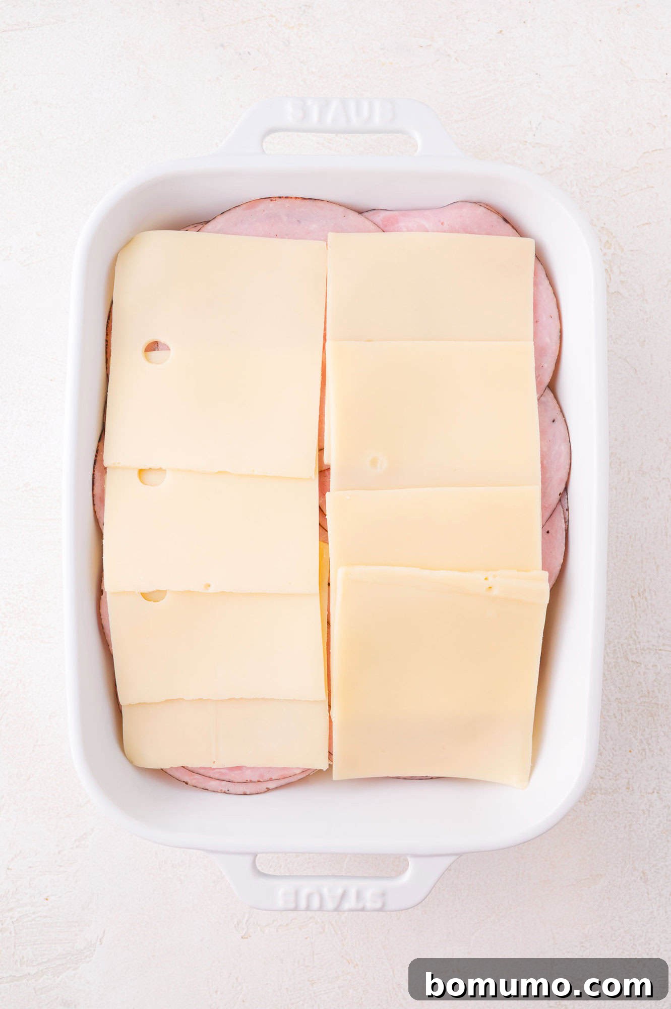 A white baking dish filled with overlapping slices of ham on the bottom and layers of Swiss cheese on top.