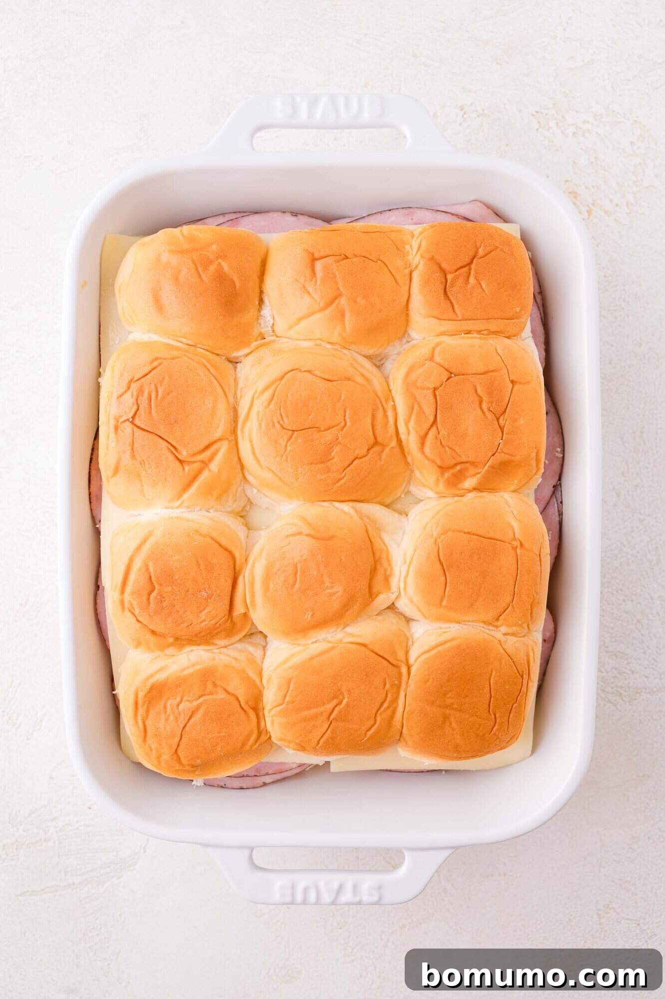 A white baking dish filled with assembled slider sandwiches, featuring a top layer of golden brown buns.