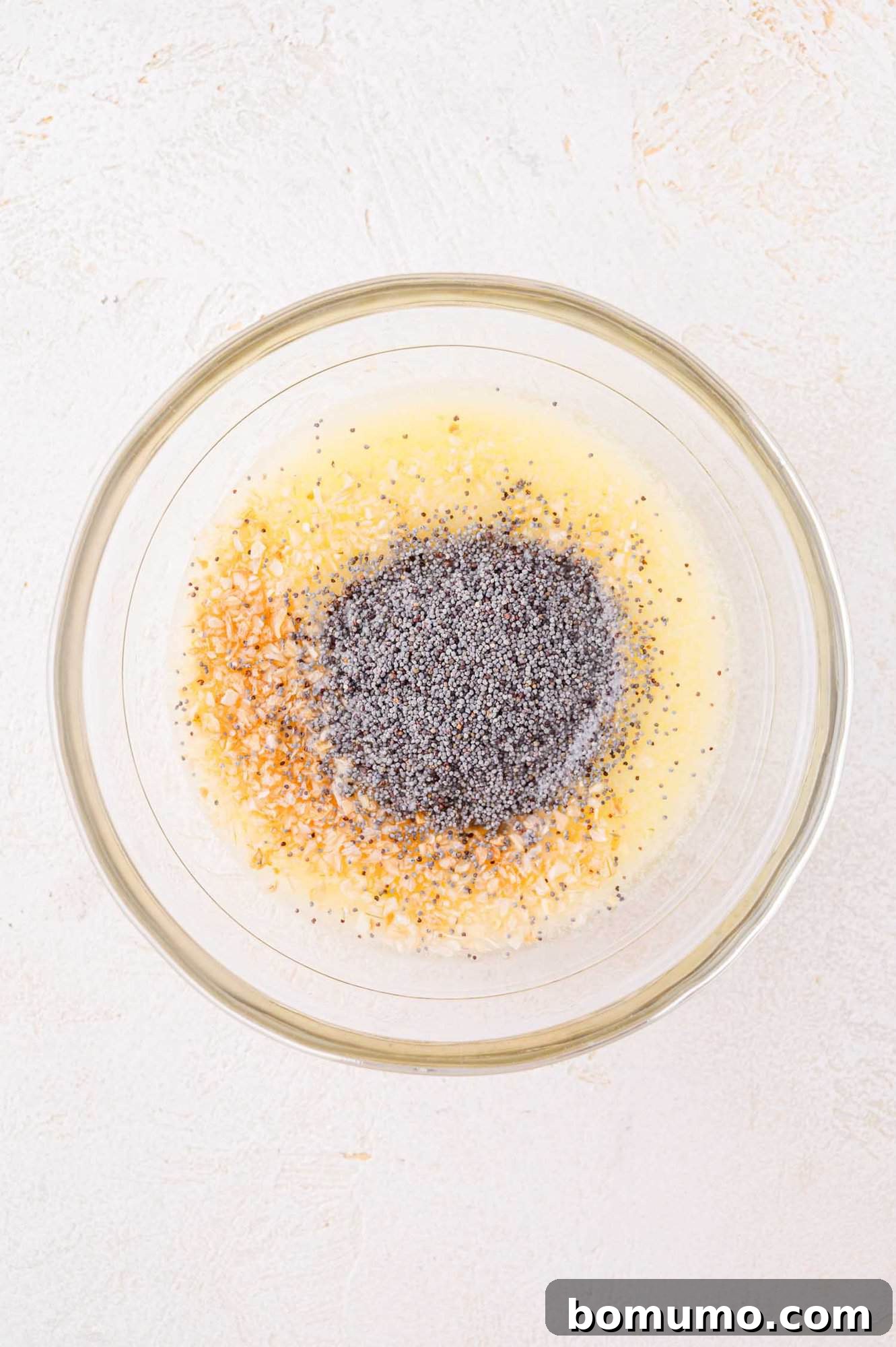 A clear glass bowl containing a mixture of poppy seeds, minced onions, and liquid ingredients on a light surface.