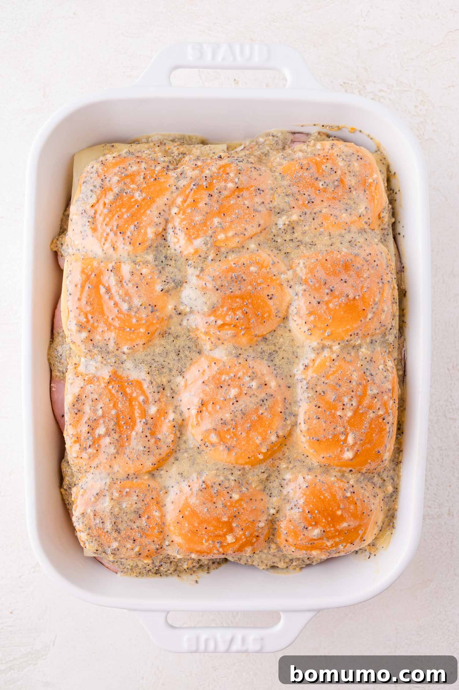 A white baking dish filled with slider sandwiches topped with a poppy seed and seasoned sauce, ready to be baked.