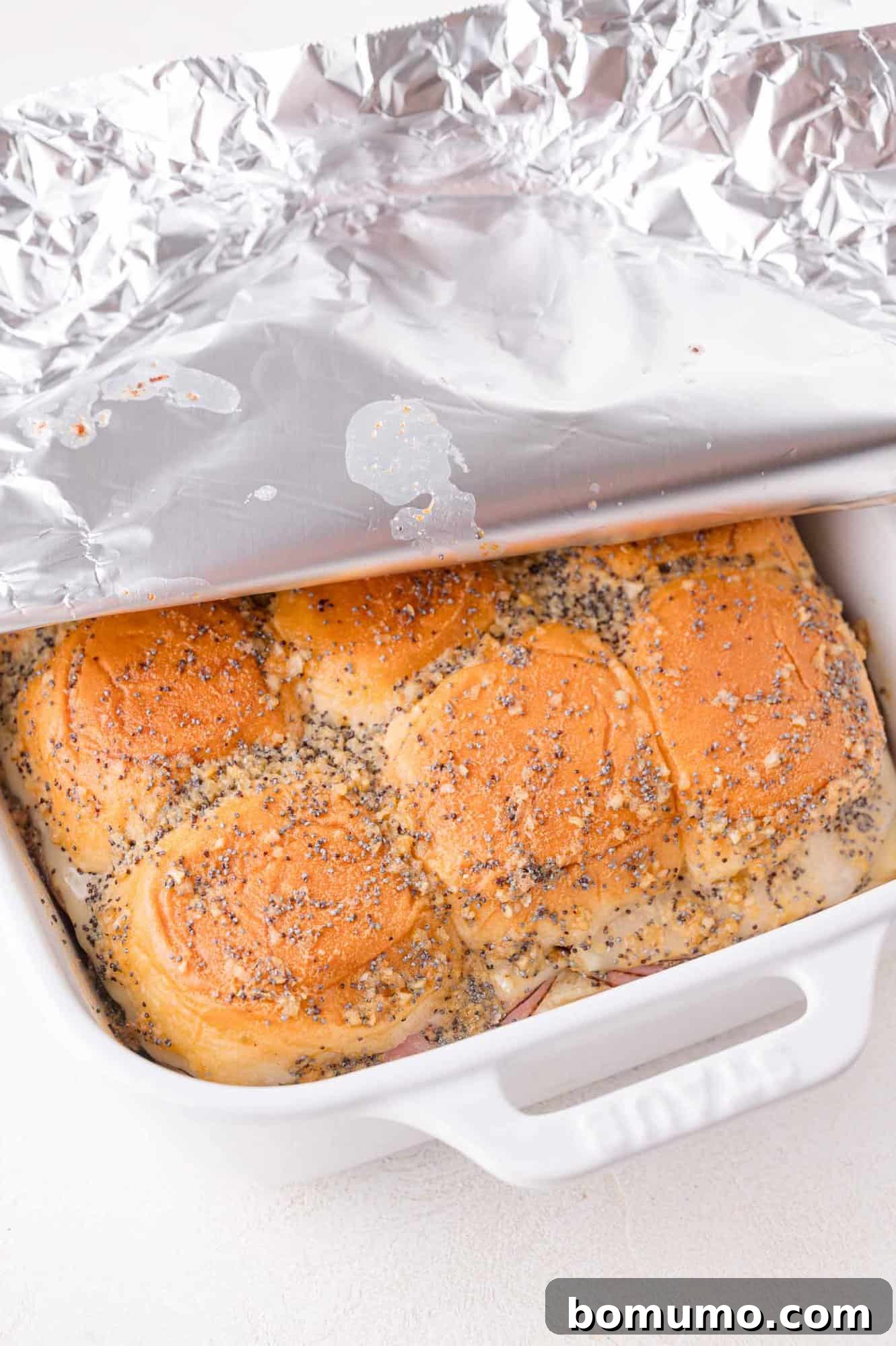 A white baking dish with poppy seed-topped buns partially covered by a sheet of aluminum foil.