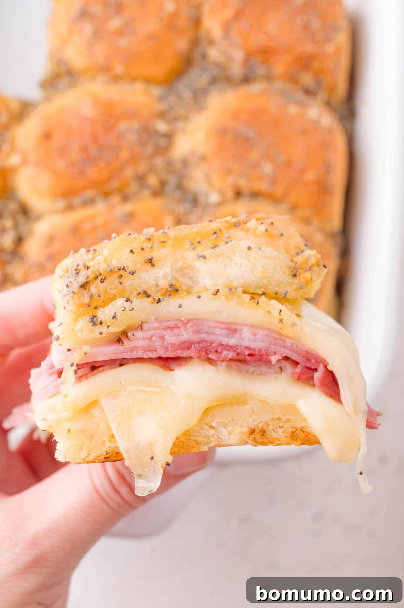 A hand holds a sandwich slider with layers of ham and melted Swiss cheese on a poppy-seed topped bun; more sliders are in the background.