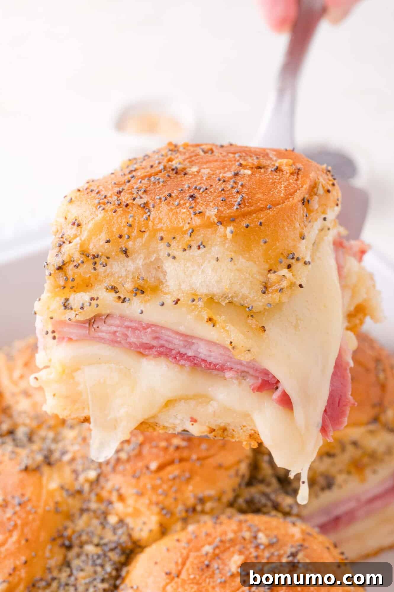 A close-up of a ham and cheese slider with poppy seeds on top, being lifted from a tray with melted cheese stretching.