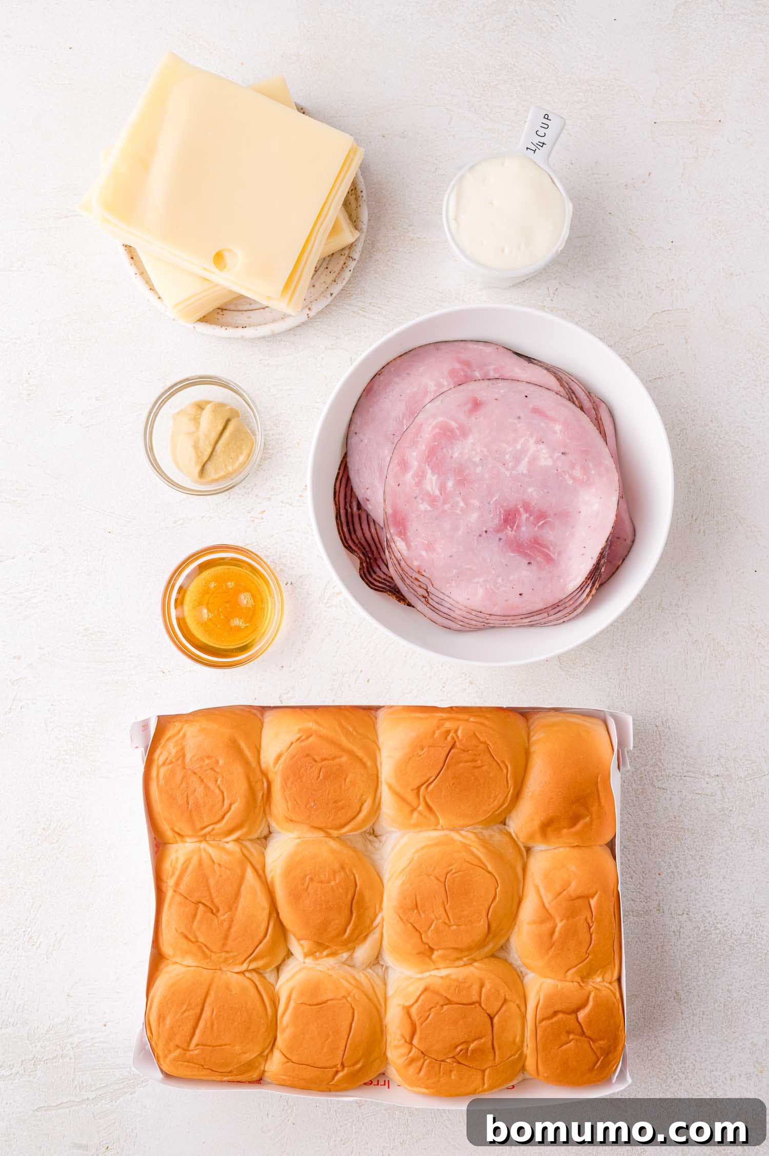 A tray of sandwich rolls next to plates of sliced ham, Swiss cheese, a bowl of mayonnaise, and small bowls of mustard and honey on a white surface.