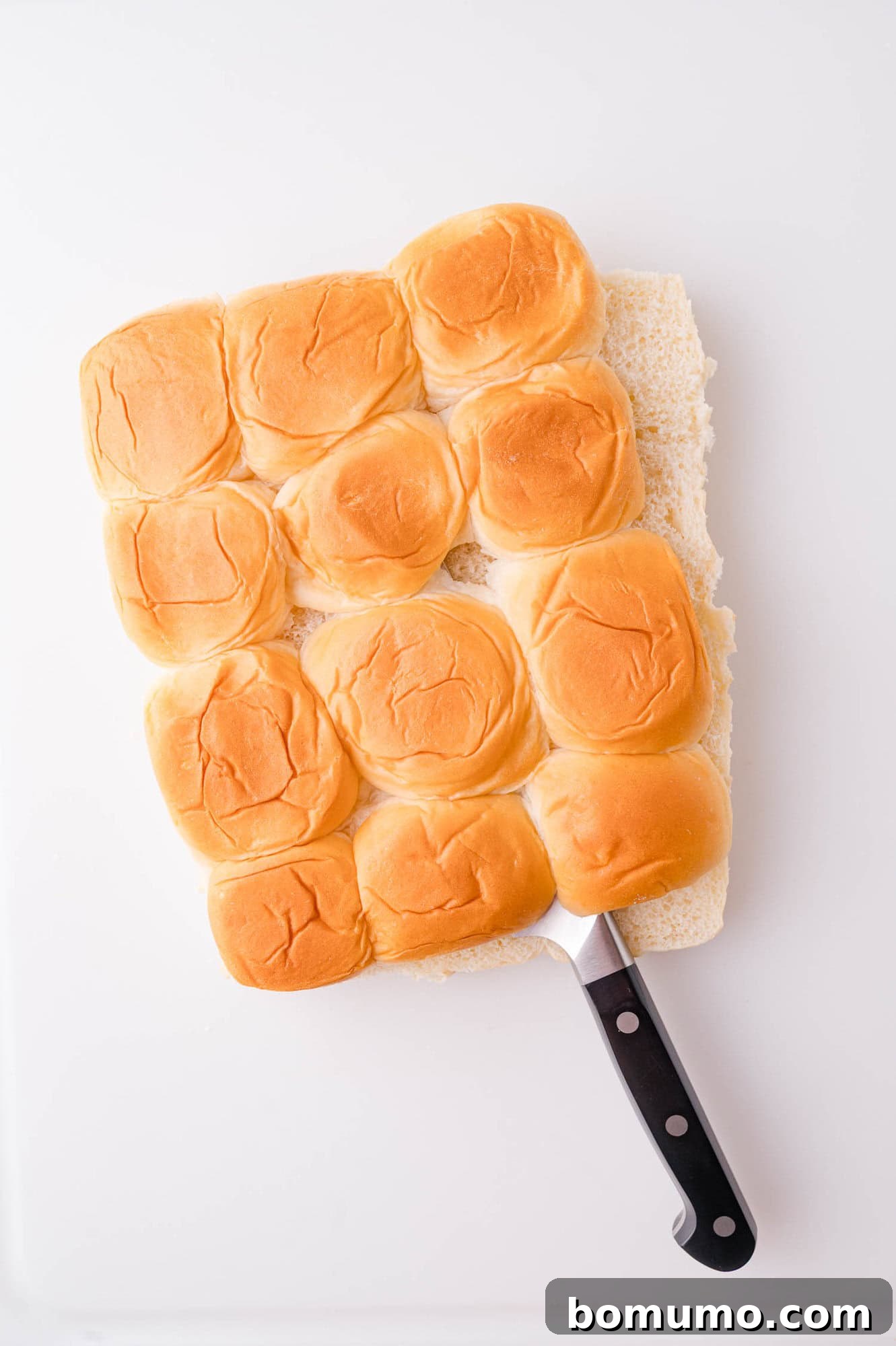 A serrated knife partially slides under a slab of twelve connected dinner rolls on a white surface.