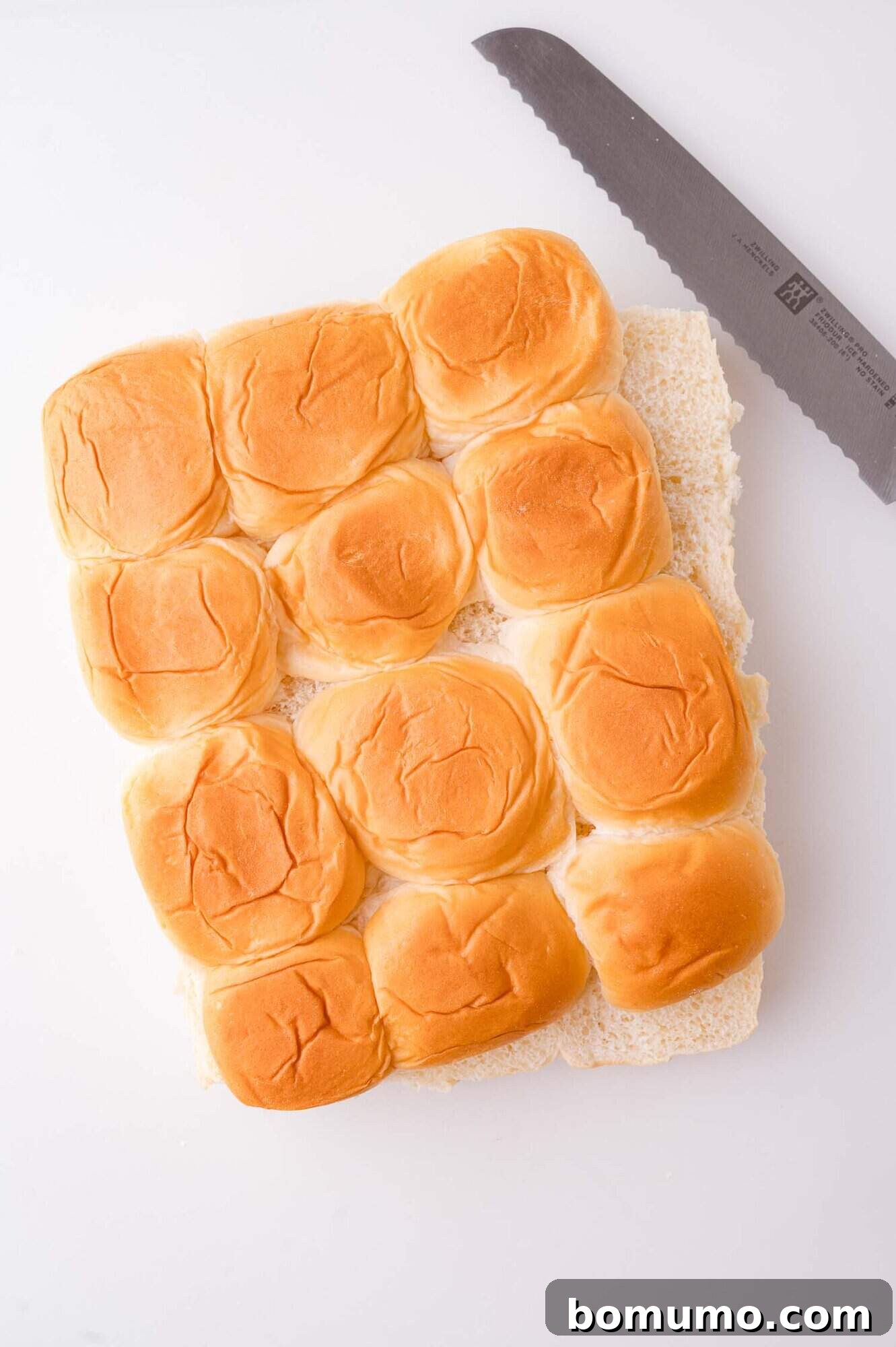 A pack of twelve sliced slider buns next to a serrated bread knife on a white surface.