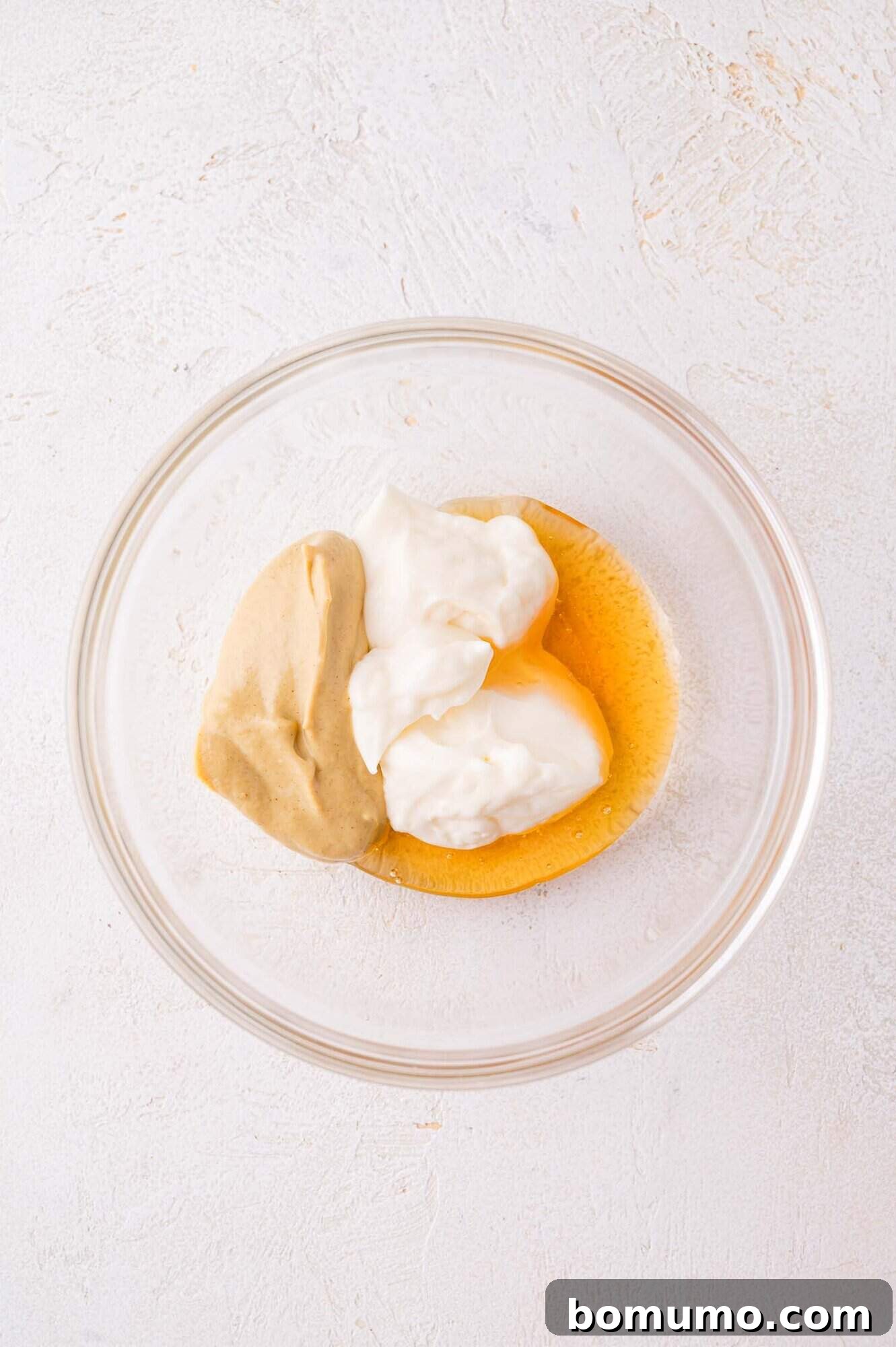 A glass bowl containing mayonnaise, mustard, and honey on a light textured surface.