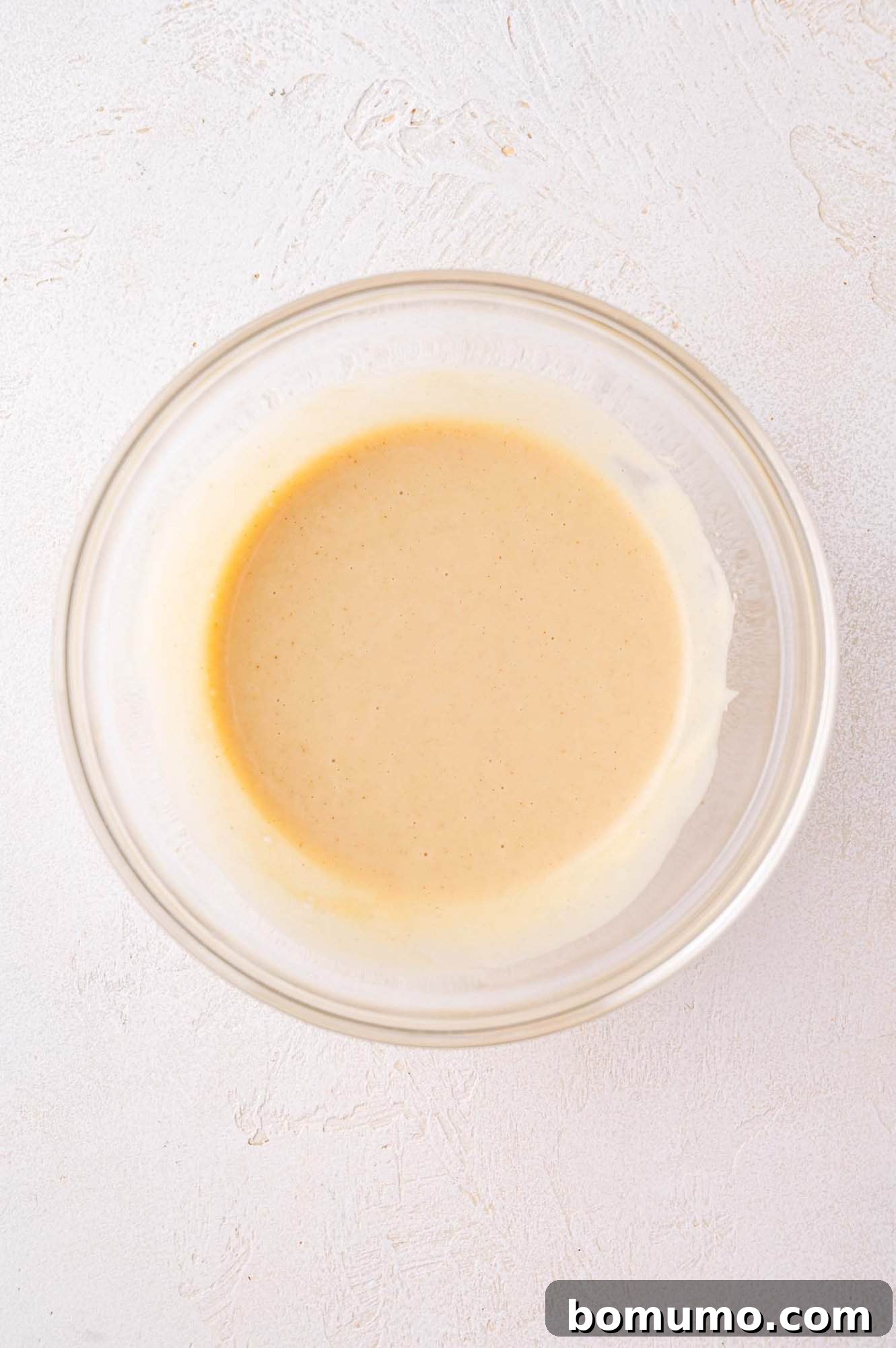 A clear glass bowl containing a light beige batter or sauce sits on a textured white surface.