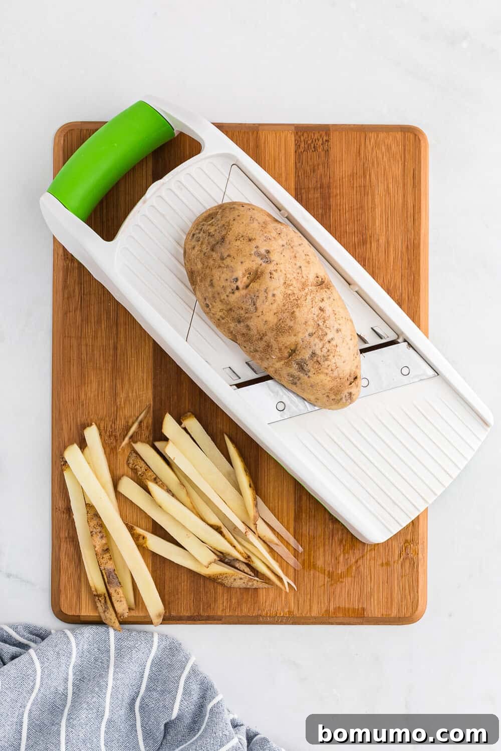 A potato being sliced on a mandoline slicer with already cut potato strips beside it, illustrating the preparation process for even fries.