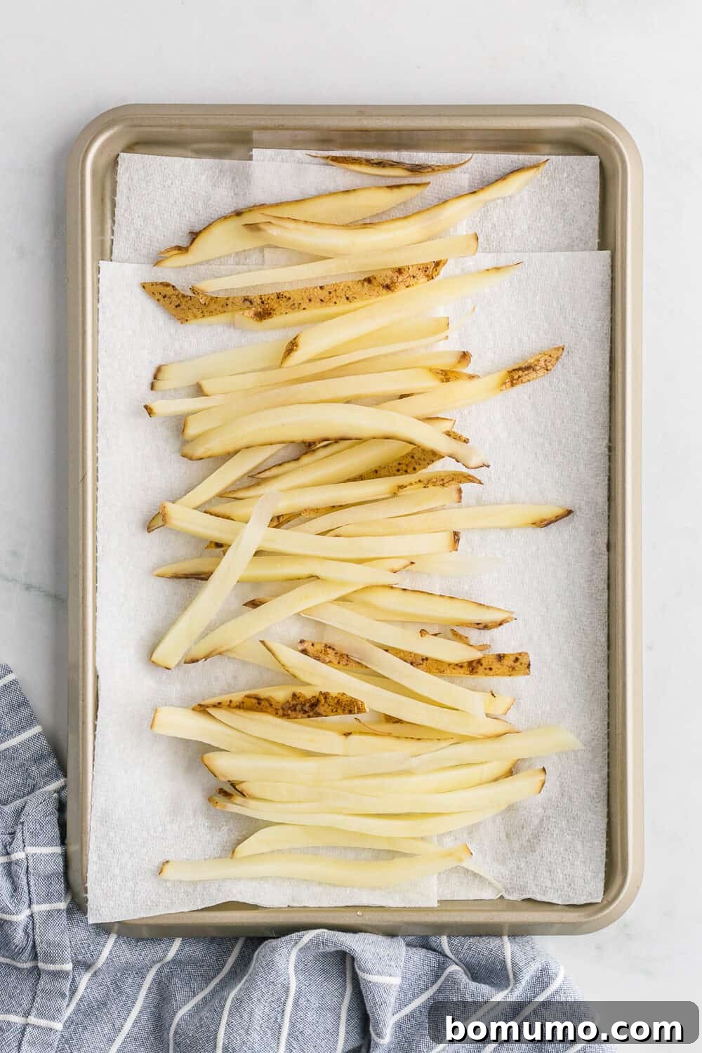 Raw potato strips spread out on a baking sheet lined with parchment paper and a striped cloth, being patted thoroughly dry before air frying.