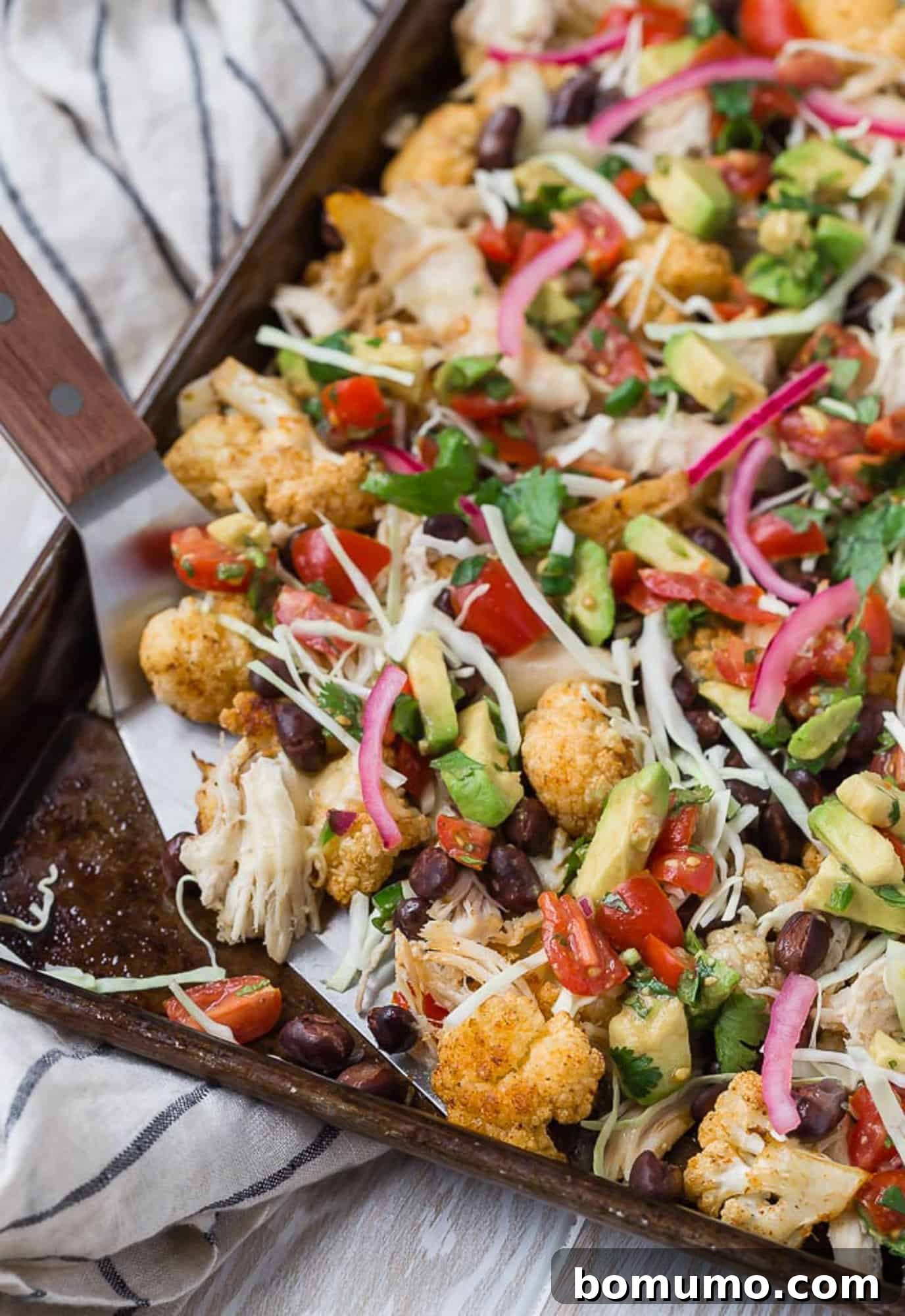 Supreme Cauliflower Nachos 2 A large serving of colorful cauliflower nachos being scooped with a spatula from a sheet pan, ready to be enjoyed.