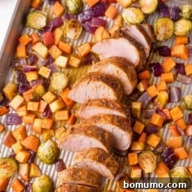 Sliced pork tenderloin with roasted Brussels sprouts, red onions, sweet potatoes, and apples on a baking sheet.