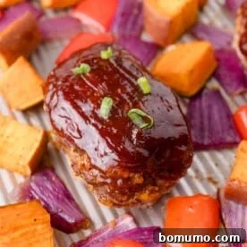 Mini meatloaf topped with barbecue sauce and green onion, surrounded by roasted sweet potatoes, red onions, and red bell peppers on a sheet pan.