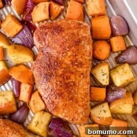 A seasoned baked pork chop on a tray surrounded by roasted sweet potatoes, apples, and red onion pieces, highlighting autumn flavors.