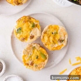 Three egg white muffins with melted cheese and visible broccoli pieces are arranged on a white plate, surrounded by ingredients and kitchen items on a light countertop.