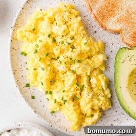Cottage cheese scrambled eggs garnished with chives, served with sliced avocado and toasted bread on a speckled plate.