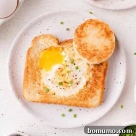 A piece of toast with a cooked egg in the center, garnished with chopped chives, served on a white plate with a toasted bread circle on the side.