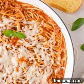 A skillet of one pan spaghetti topped with melted cheese, garnished with basil, sitting on a light surface with a slice of bread nearby.