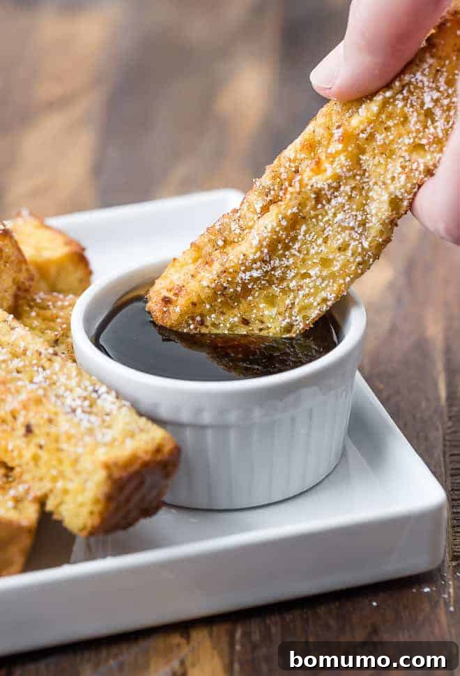 A hand dipping a French toast stick into a bowl of maple syrup on a plate, next to more French toast sticks.