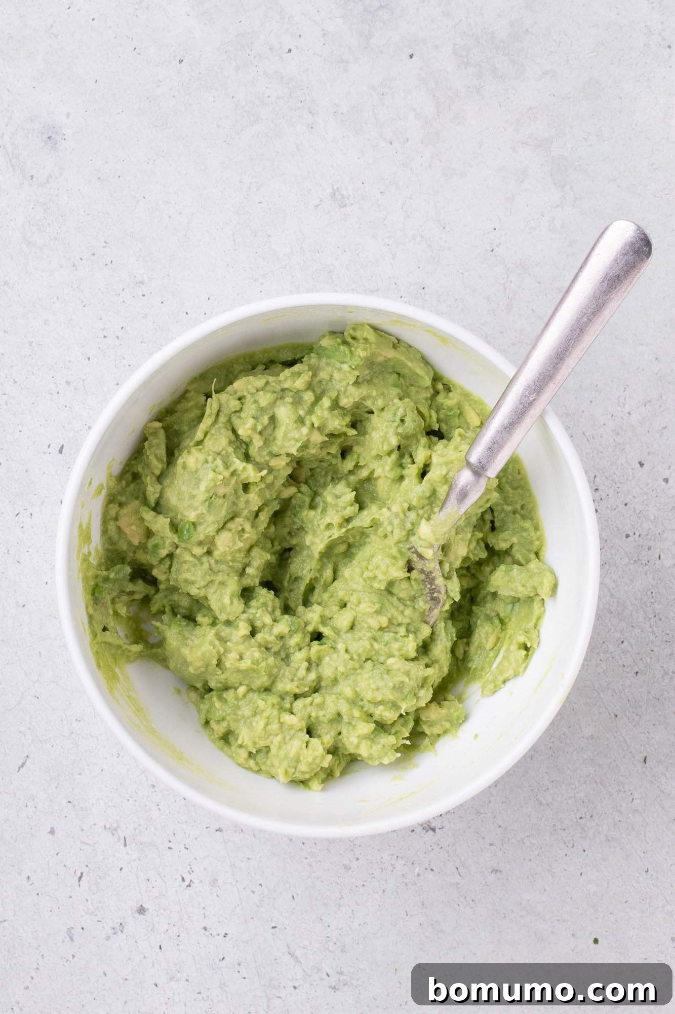 Bowl of mashed guacamole with a spoon on a light gray surface.