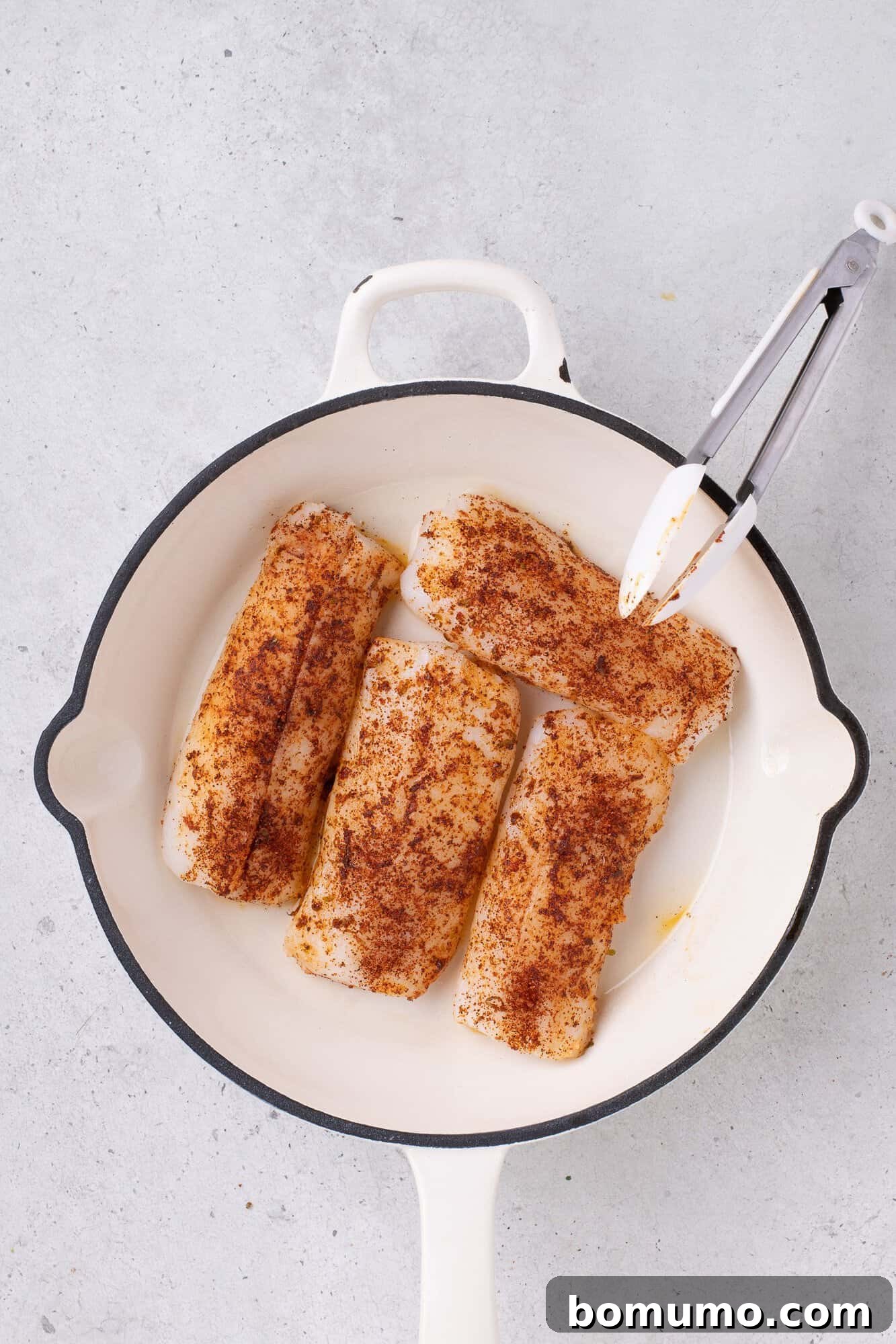 Four seasoned fish fillets in a white frying pan with tongs, placed on a light gray surface.