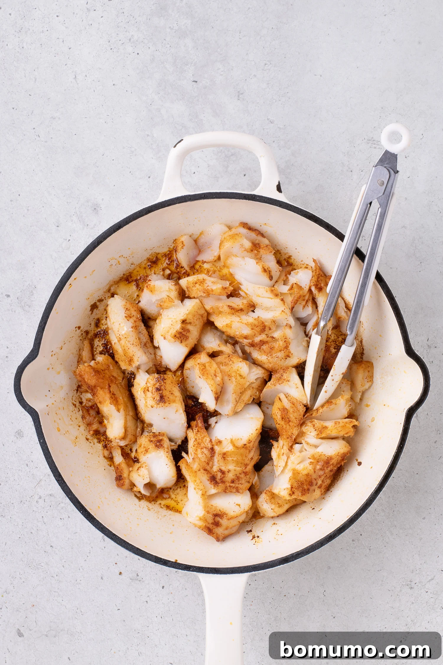 Cooked pieces of seasoned fish in a white skillet with metal tongs on a light surface.