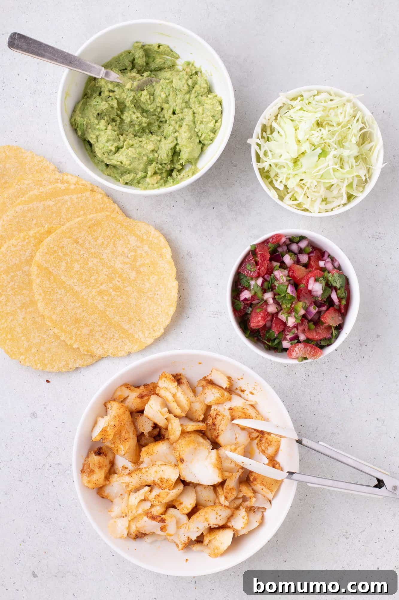 Ingredients for fish tacos on a table: corn tortillas, guacamole, shredded cabbage, salsa, and seasoned fish pieces in separate bowls.