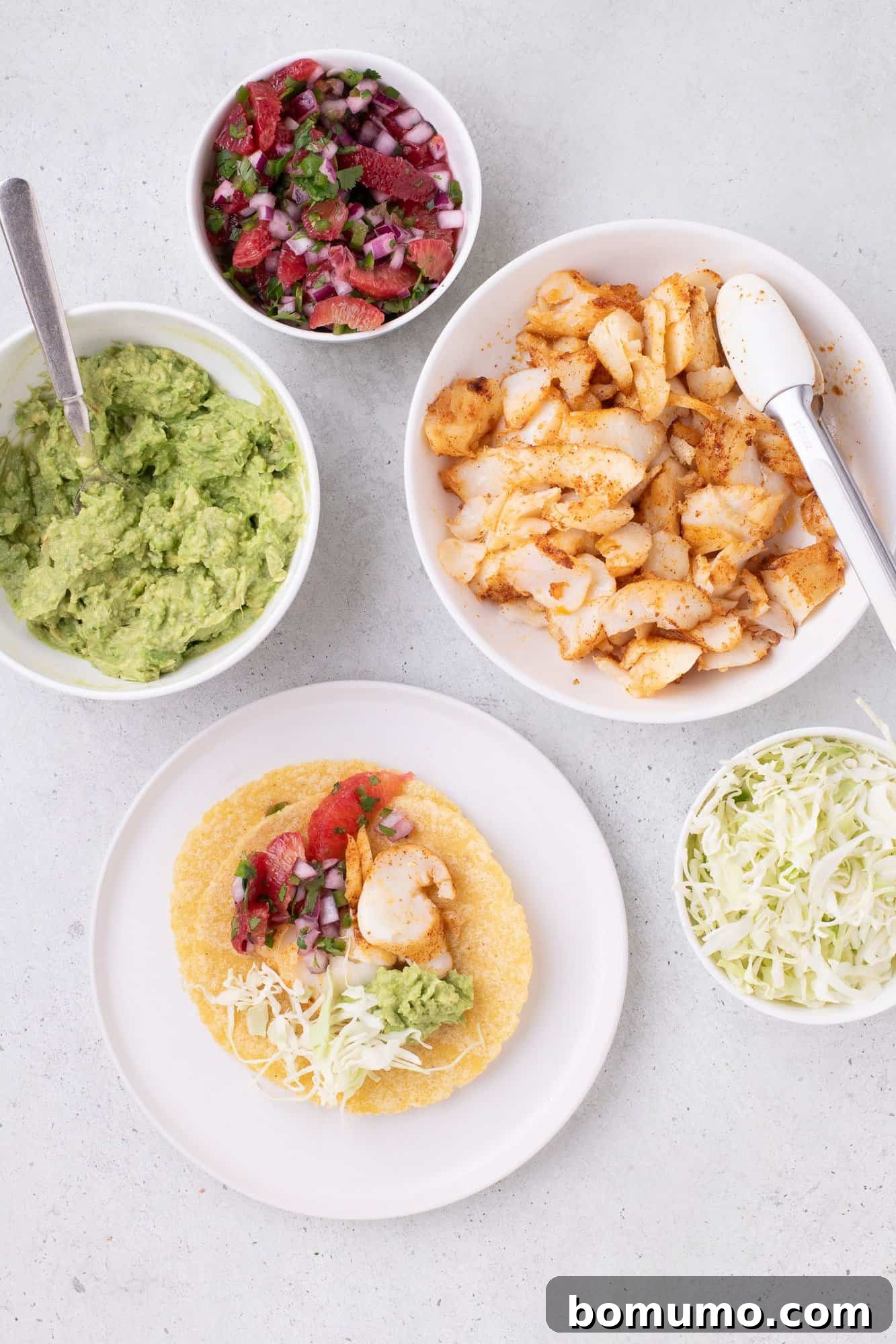Taco ingredients with corn tortillas, seasoned fish, guacamole, cabbage, and salsa displayed on a light surface.