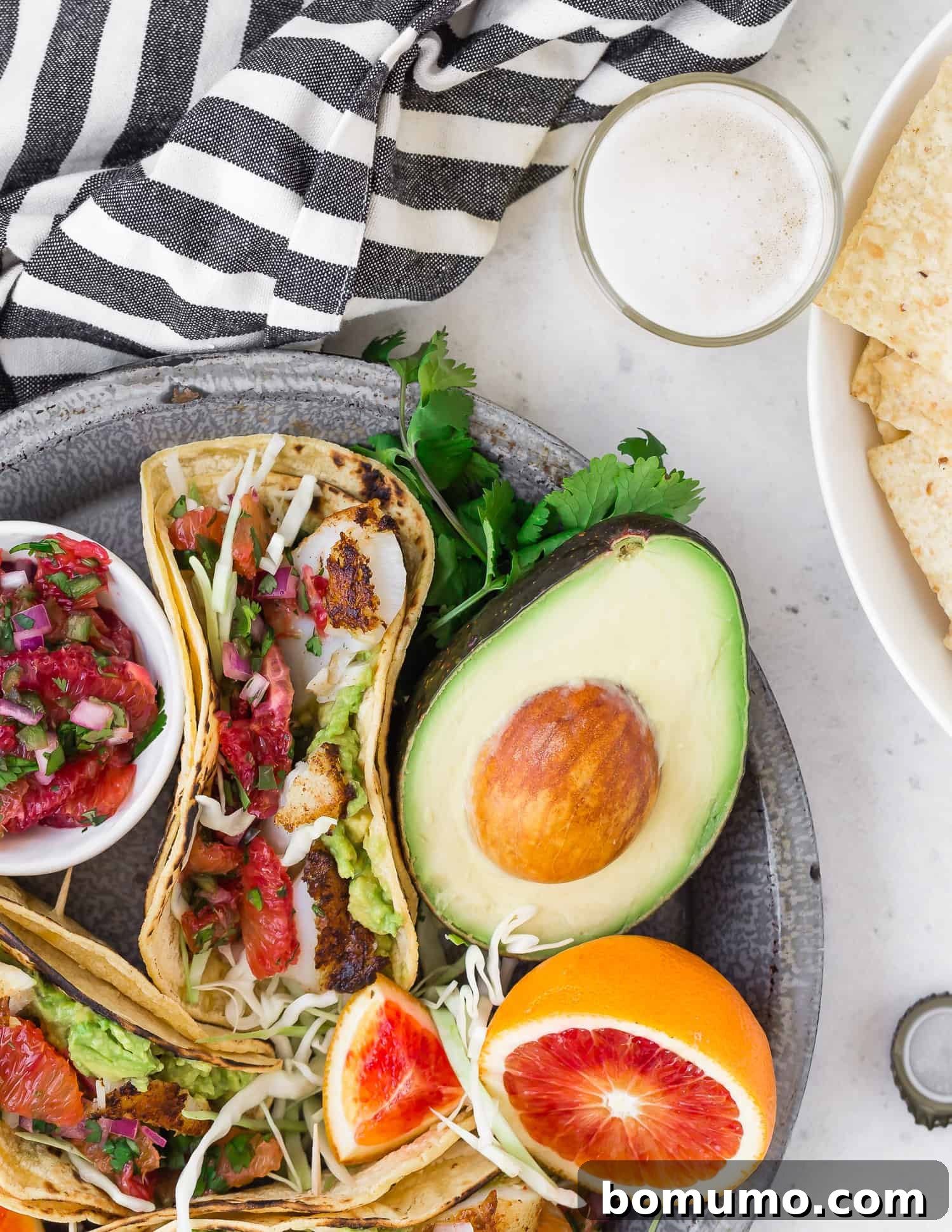 Tacos on a platter with avocado, blood orange, a beer, and chips nearby.