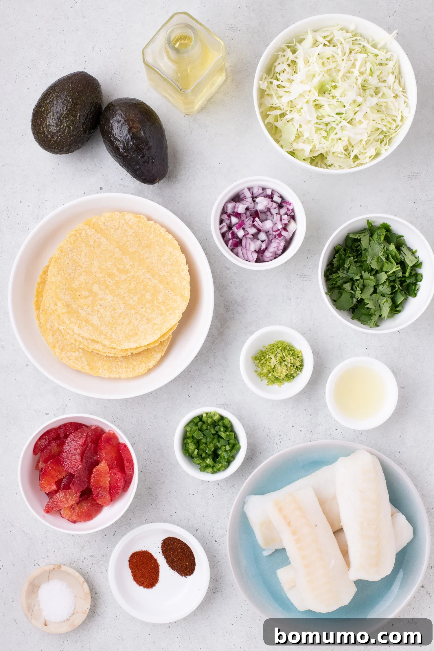 Ingredients for fish tacos laid out: corn tortillas, fish fillets, sliced cabbage, avocado, lime zest, lime juice, red onion, cilantro, jalapeño, chili powder, and salt.