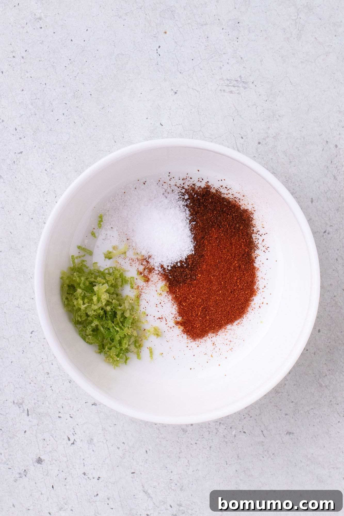A bowl with salt, paprika, and lime zest on a light textured surface.