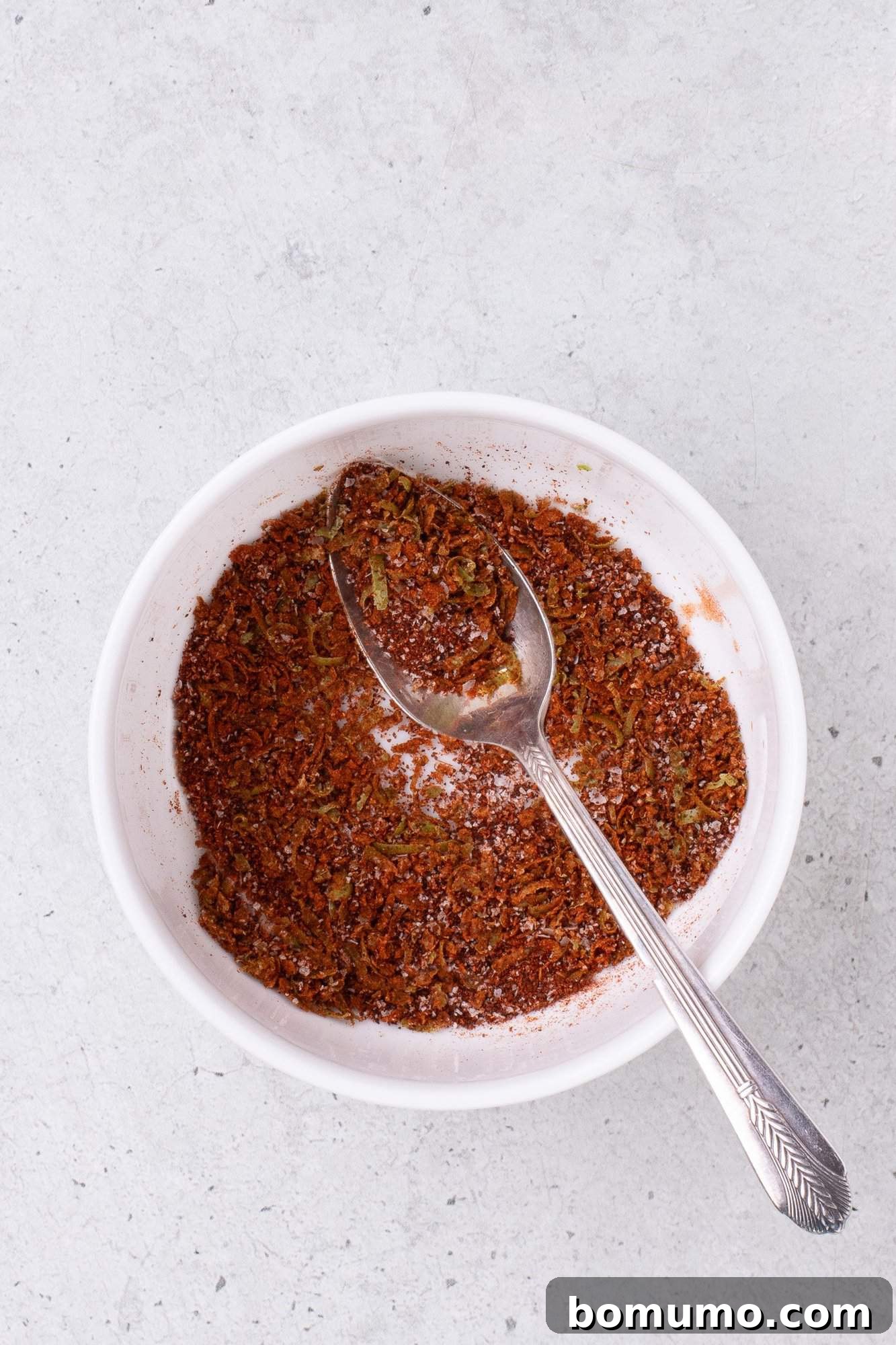A bowl containing a mixture of red spices and herbs with a spoon resting inside.