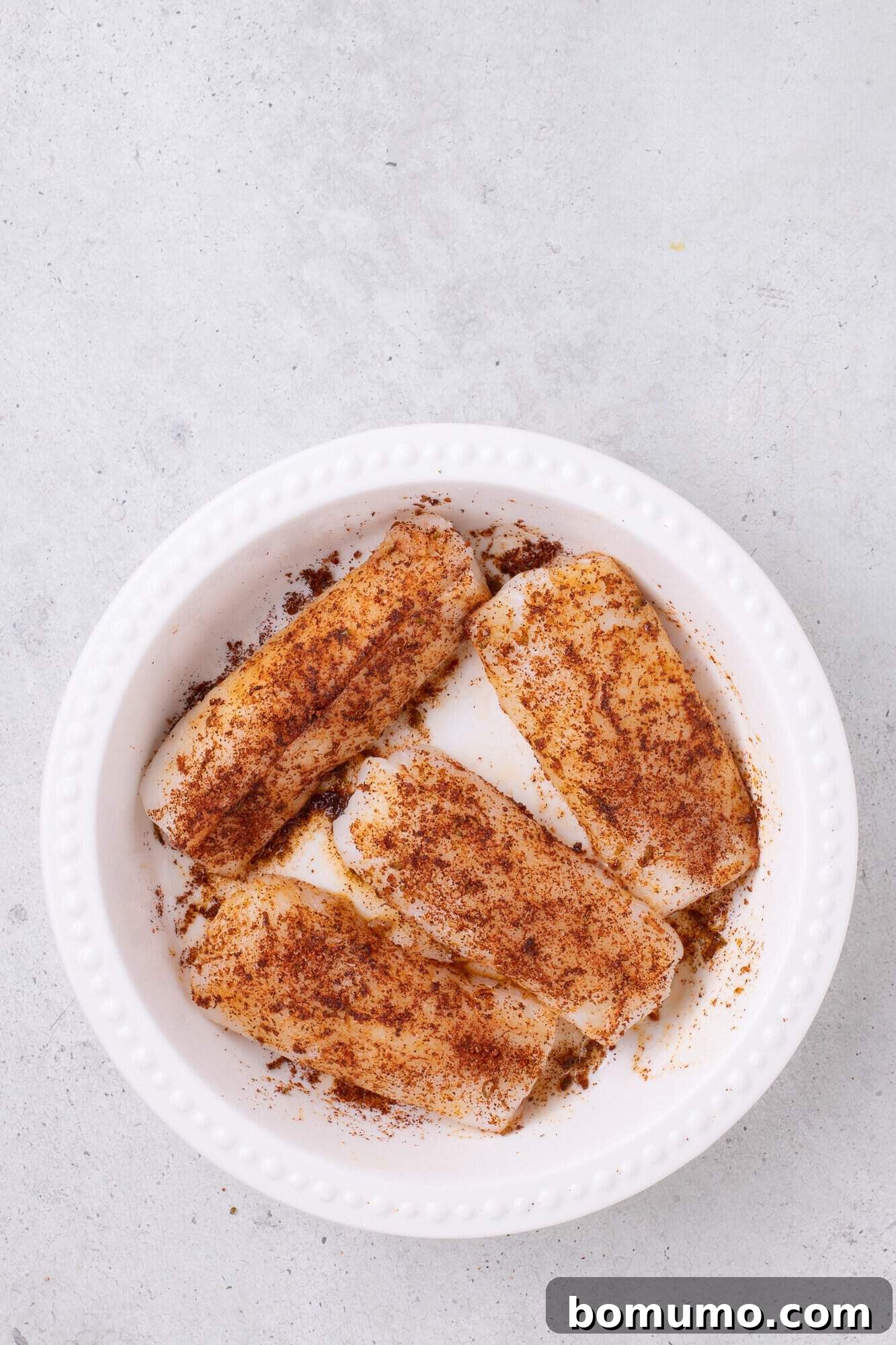 Four raw fish fillets in a white dish, sprinkled with seasoning on a light gray background.