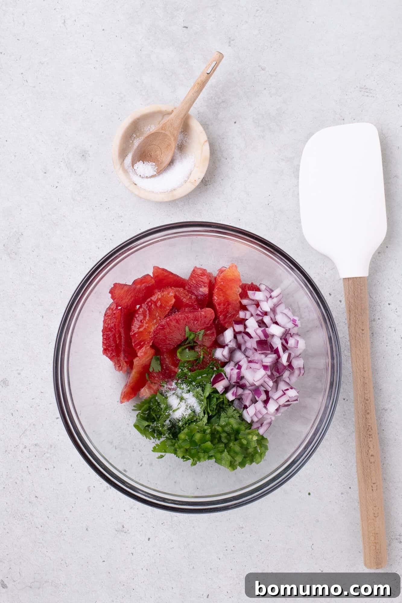 Glass bowl with diced tomatoes, red onion, jalapeño, and cilantro. Nearby are a small wooden bowl with salt and a white spatula on a gray surface.