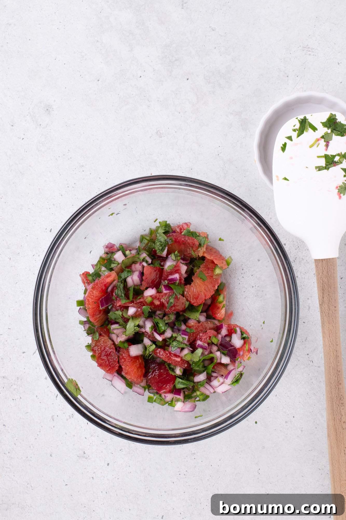 A bowl of diced red onions, cilantro, and grapefruit segments, with a spatula nearby on a light-colored surface.
