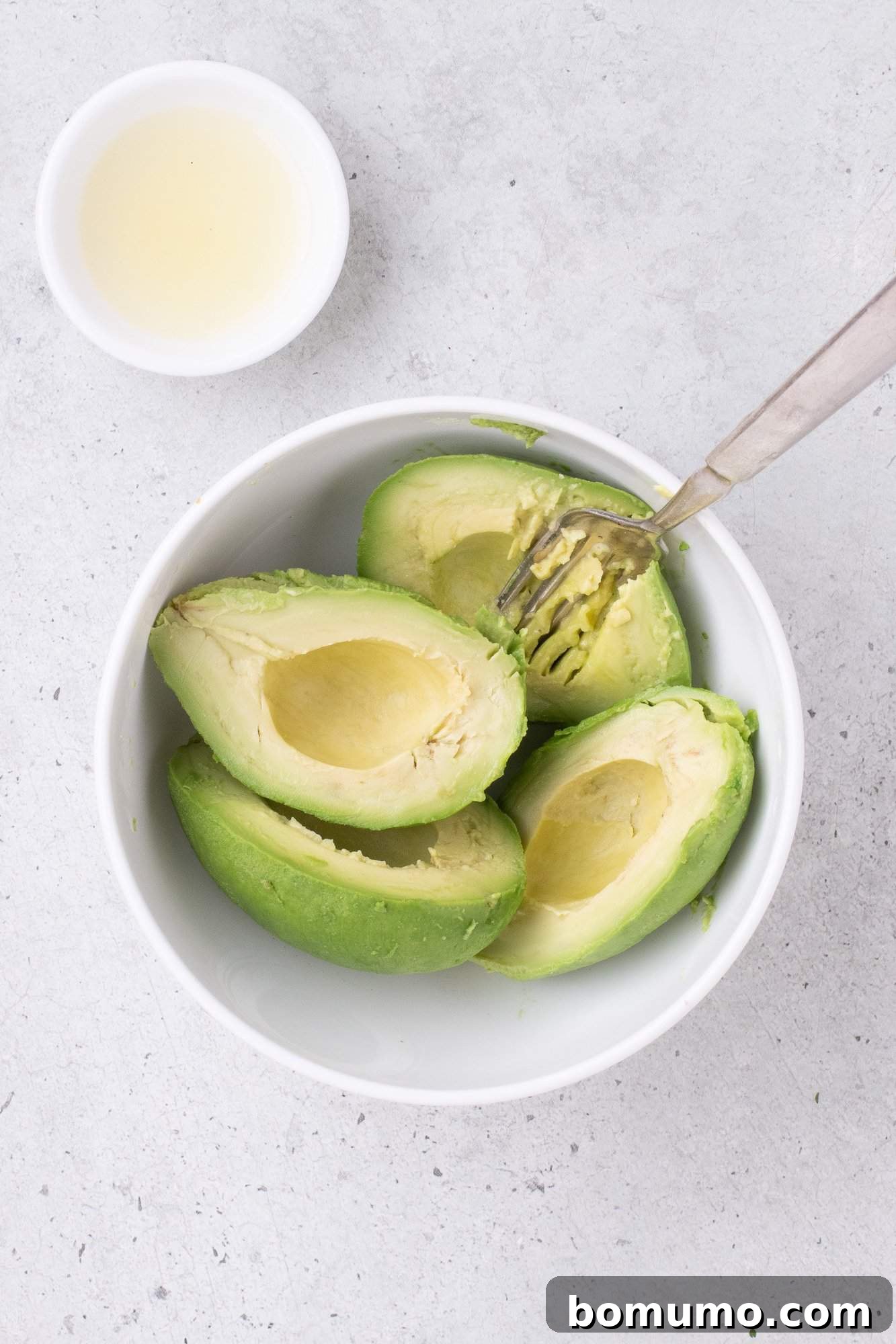 A bowl with four avocado halves and a fork next to a small bowl of lemon juice on a light gray surface.