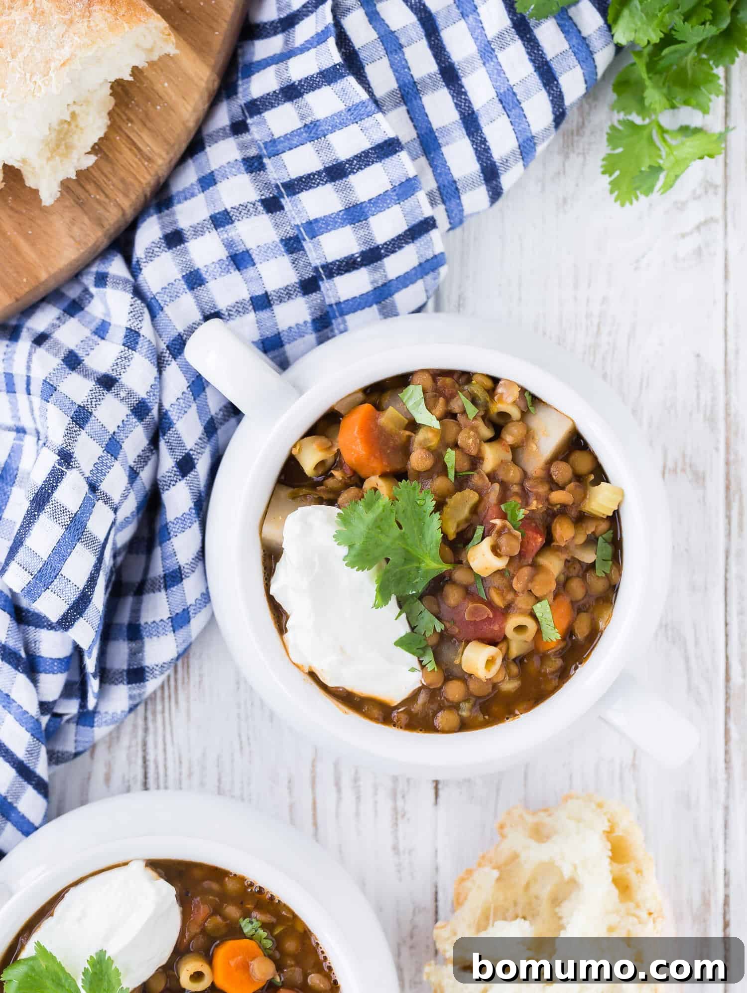 Overhead photo of two bowls of lentil soup, one only partially showing in image. Cilantro is also pictured as well as fresh bread that has been torn from the loaf, ready for dipping.