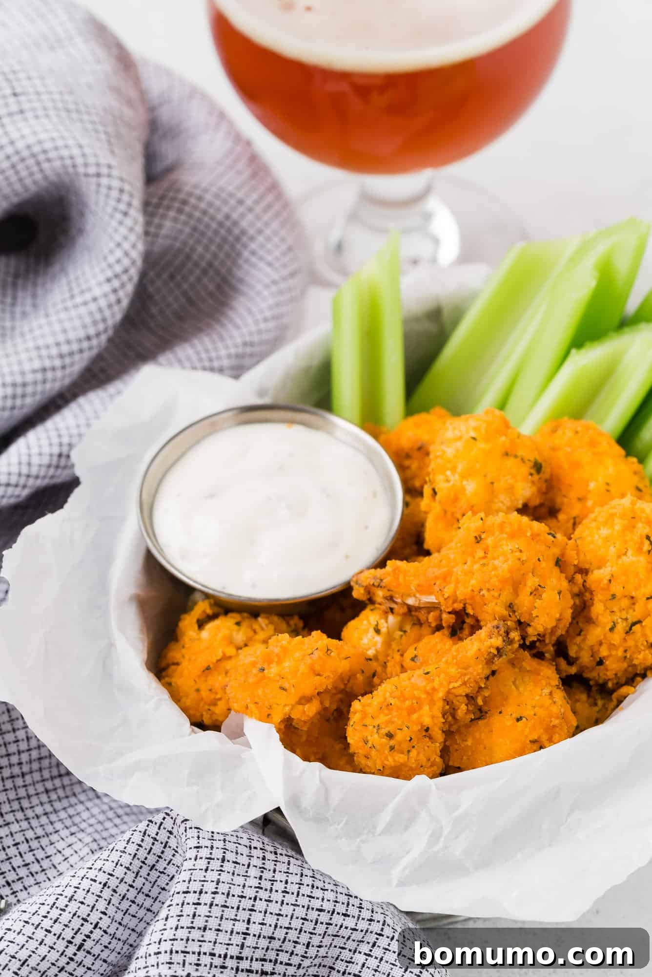 Crispy buffalo cauliflower bites in a metal basket, with a blurred background of a frothy glass of beer, setting a perfect casual dining scene.