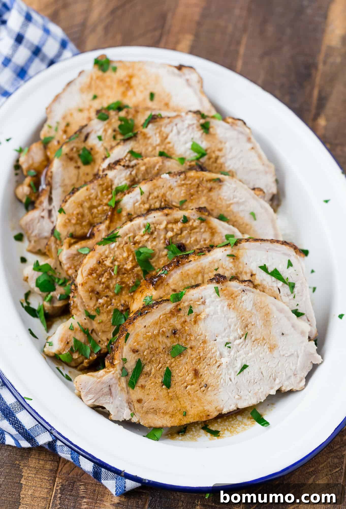 Sliced roasted pork served on a white oval platter, garnished with chopped herbs. A blue and white checkered cloth is partially visible beside the platter.