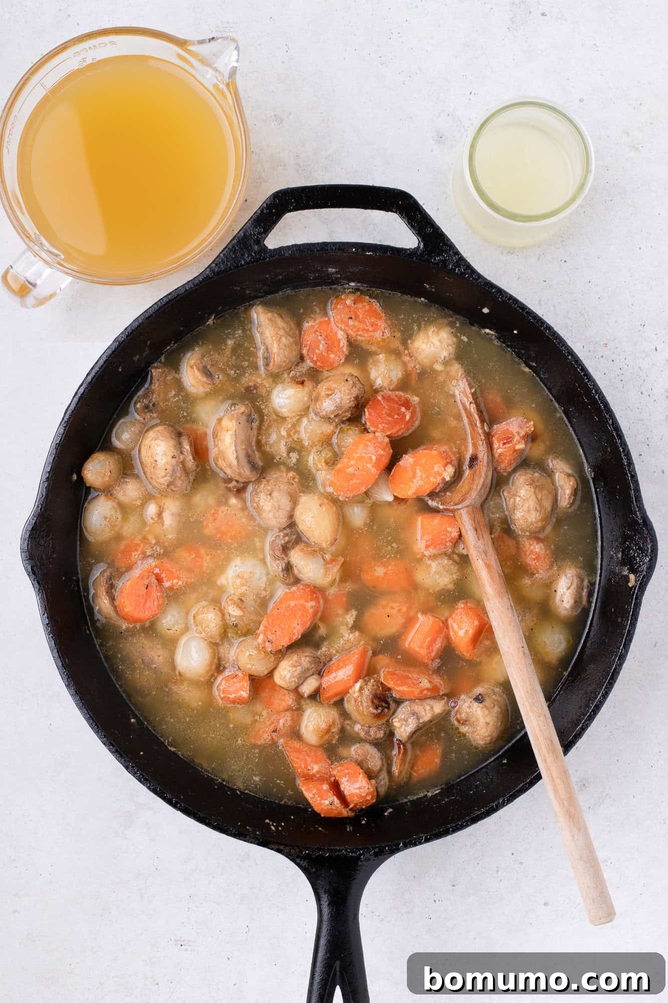 Chicken broth being poured into the skillet with sautéed vegetables and roux, forming the base of the fricassee sauce.