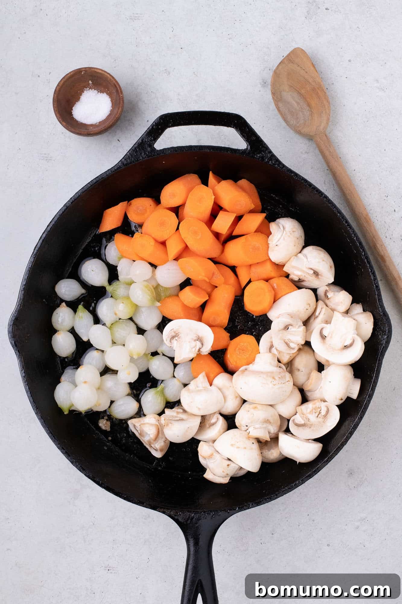 Freshly cut vegetables, including carrots, mushrooms, and pearl onions, waiting in the skillet before cooking.