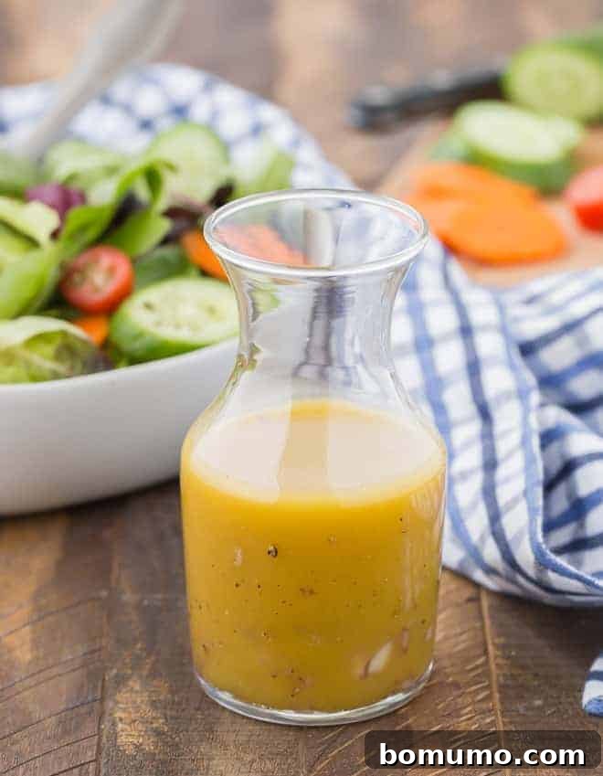 A small jar of homemade red wine vinaigrette sits on a wooden surface, with a fresh tossed salad and chopped vegetables blurred in the background, suggesting a healthy meal.