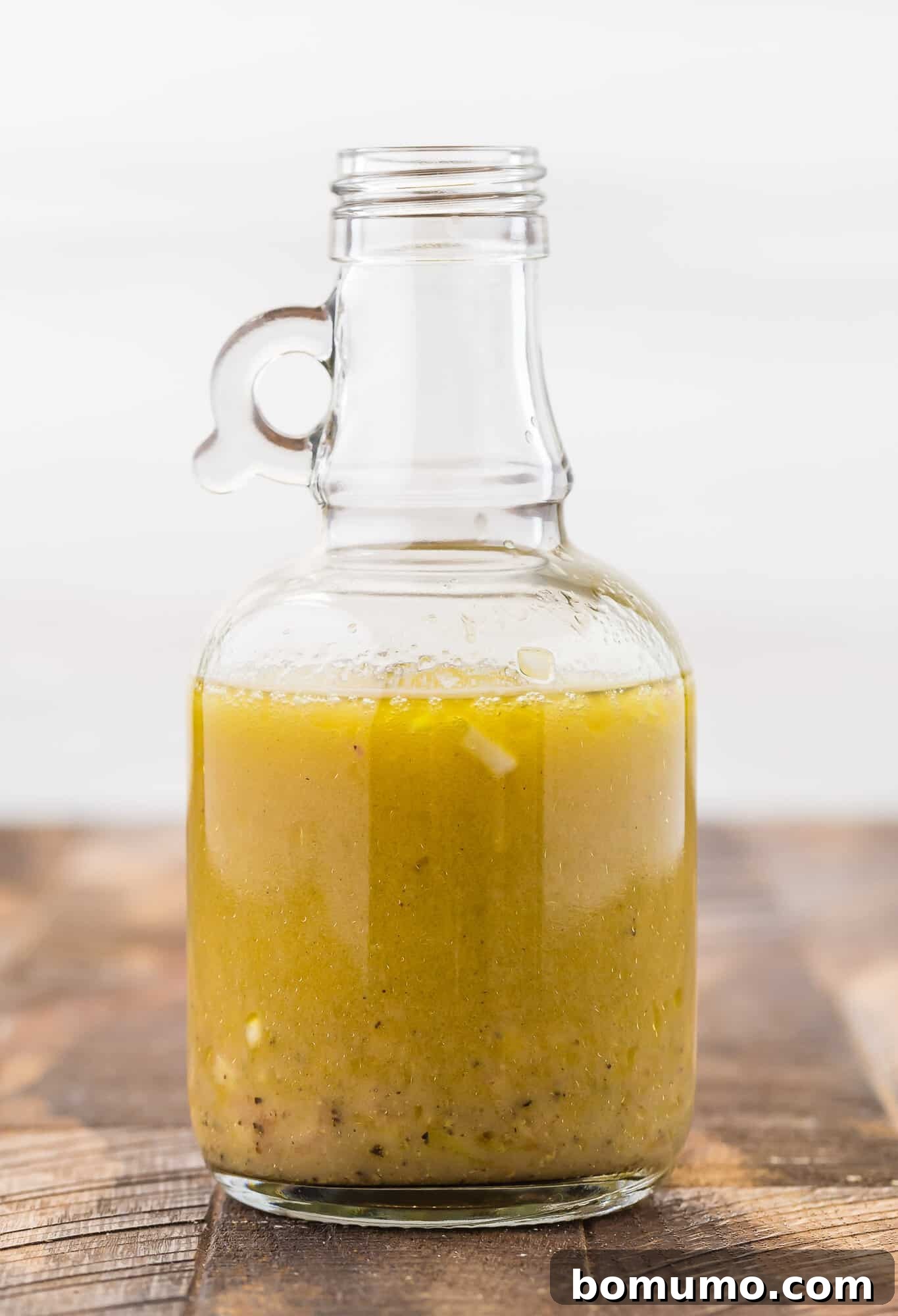 A small glass jar of golden white wine vinaigrette sits on a light wooden table, with fresh green ingredients blurred in the background, highlighting the homemade dressing.