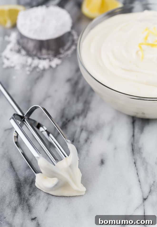 Image of lemon cream cheese frosting in a glass bowl. A beater is next to it, with frosting on it. In the background are powdered sugar and lemons.