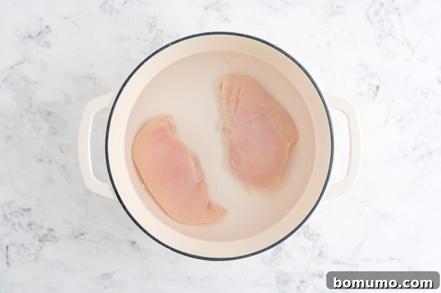 Raw chicken breasts fully covered in water within a pan, preparing for poaching.